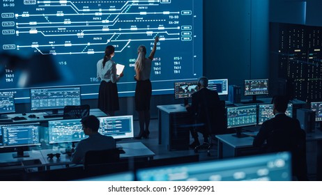 Two Software Engineers Talking Next to a Big Screen in a Modern Monitoring Office with Live Analysis Feed with Charts. Monitoring Room Big Data Scientists and Managers Sit in Front of Computers.