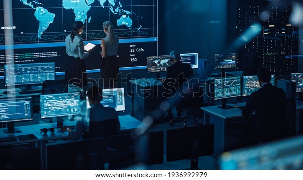 Two Software Engineers Talking Next to a Big\
Screen in a Modern Monitoring Office with Live Analysis Feed with\
Charts. Monitoring Room Big Data Scientists and Managers Sit in\
Front of Computers.