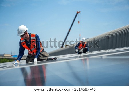 Two solar installation engineers in white helmets and high visibility vests deliberately kneel together at the array edge preparing the next fixings on the curved metal roof.