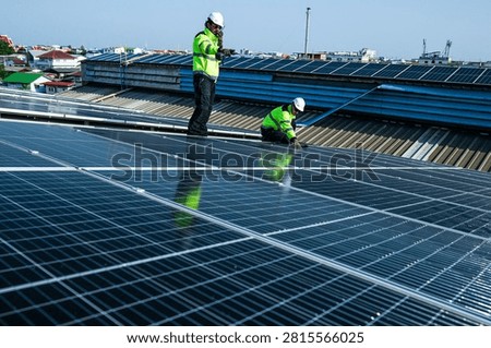 Two solar installation engineers in white helmets and high visibility vests skilfully crouch inspecting the huge array seen from high above on the curved metal roof of an industrial factory.