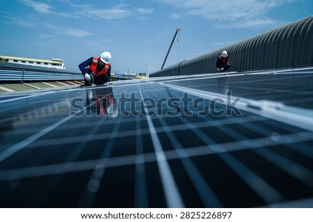 Two solar installation engineers in white helmets and high visibility vests skilfully kneel fastening clamps against a towering monsoon sky on the curved metal roof of an industrial factory.