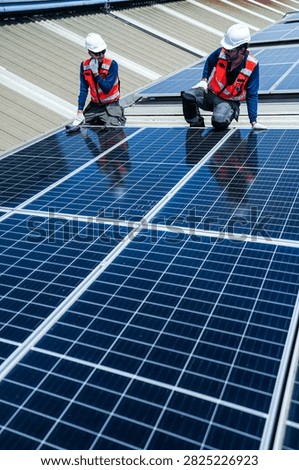 Two solar installation engineers in white helmets and high visibility vests skilfully move across the giant module grid checking row by row on the curved metal roof of an industrial factory.