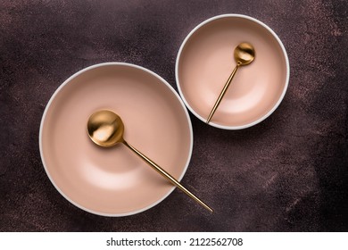 Two soup bowls and spoons on old dark brown table with scuffs. View from above. Dishes and utensils for serving and eating meals. Beige round rimmed plates and gold colored cutlery. Modern crockery.