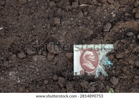 Two Soviet-era 100-ruble notes emerge from the soil, representing a forgotten past. Once valuable currency of a superpower, they now rest in the earth as symbols of a fallen state, consigned to the pages of history.