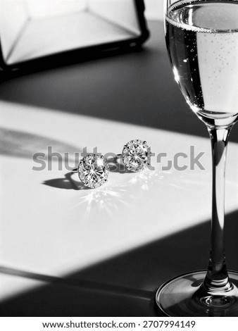 Two sparkling diamond stud earrings with halo settings are placed next to a tall, bubbly glass of champagne in a dramatic black and white image.