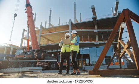 Two Specialists Inspect Commercial, Industrial Building Construction Site. Real Estate Project with Civil Engineer, Investor Use Laptop. In the Background Crane, Skyscraper Concrete Formwork Frames