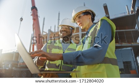 Two Specialists Inspect Commercial, Industrial Building Construction Site. Real Estate Project with Civil Engineer, Investor Use Laptop. In the Background Crane, Skyscraper Concrete Formwork Frames