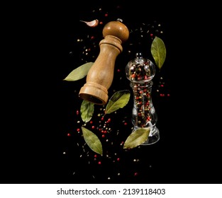 Two spice mills with peppercorns and bay leaves flying over black background. Grinder for spices. Salt and pepper shakers levitation