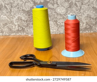 Two spools of thread in yellow and red sit next to a pair of scissors on a wooden table. The scene shows tools used for sewing or crafting activities indoors.