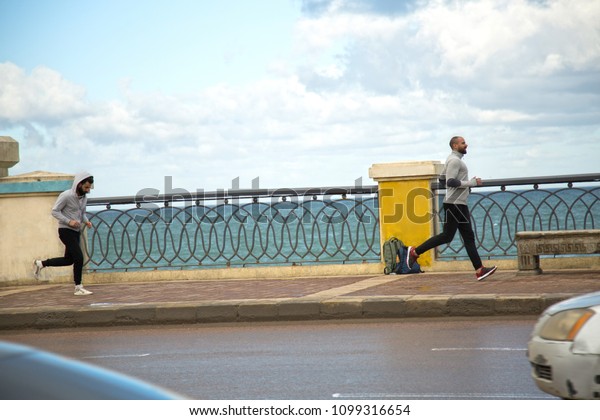 Two sportive friends\
jogging on the sidewalk by the sea one of them look tired running\
behind his friend