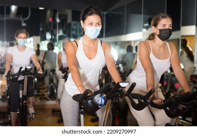 Two Sportive Girl In Face Masks Riding Exercise Bike During Cycling Class In Gym