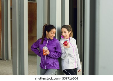 Two Sporty Female Friends Taking A Fitness Workout Rest For A Healthy Snack Eating A Red Apple And Drinking Orange Juice Or Fruit Detox Smoothie.