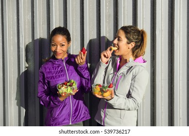 Two Sporty Female Friends Taking A Fitness Workout Rest For A Healthy Snack With Fruits And Green Salad.