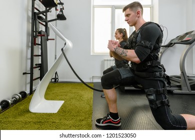 Two sporty people in EMS costumes squatting in modern gym