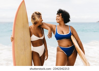 Two sporty women enjoying themselves in their beach life, standing on shore with surfboards in hand and happy smiles on their faces. Female surfers embarking on a surfing adventure in bikinis. - Powered by Shutterstock