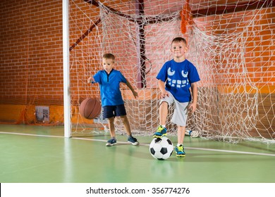 Two Sporty Young Boys On An Indoor Court Playing In The Goalposts, One Bouncing A Basketball And The Other Standing On A Soccer Ball