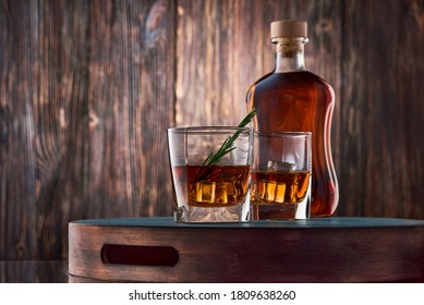 Two square glasses of whiskey with ice and a sprig of rosemary, and a full bottle of whiskey, stand on a tray against an old wooden wall. Low key.