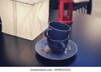 Two stacked dark blue teacups on a saucer with a white lamp and red lantern on a black table. Indoor home decor and tableware concept for posters and prints. - Powered by Shutterstock