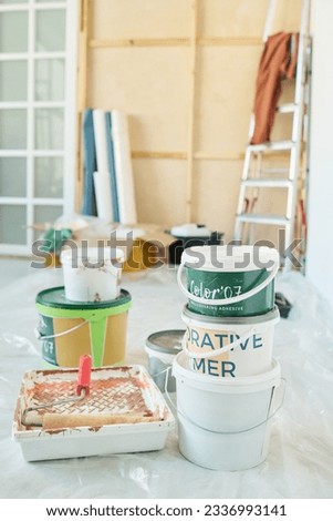 Two stacks of plastic buckets containing paints for walls and paintroller in tray standing on the floor of living room in new apartment