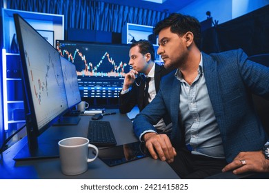 Two stock exchange traders discussing dynamic investment graph in currency rate on monitor at night. Businessman partners coffee meeting in high stock market in neon light at workplace. Sellable. - Powered by Shutterstock