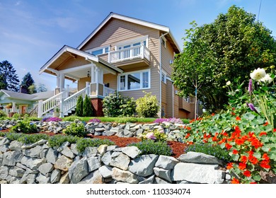 Two story beige nice house on the hill with rock walls and flowers.