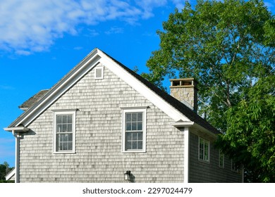 Two story grey colored vintage building with wood cedar shake siding, two double hung windows, and a chimney. There's a large lush green tree taller than the house. The trim on the building is white.