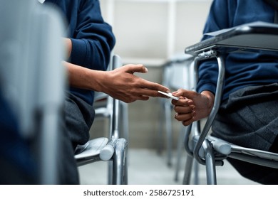 Two students in a classroom passing a note discreetly. Hands exchanging a note, seated at desks. Classroom setting, note passing, student interaction. Students cheating on an exam, passing a note.