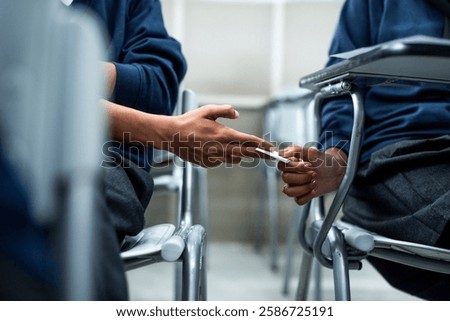 Two students in a classroom passing a note discreetly. Hands exchanging a note, seated at desks. Classroom setting, note passing, student interaction. Students cheating on an exam, passing a note.