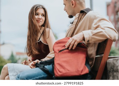 Two students sit on a bench outdoors, talking and smiling. A young man and woman converse casually with a backpack between them.