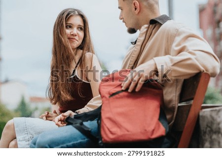 Two students sit on a bench outdoors, talking and smiling. A young man and woman converse casually with a backpack between them.