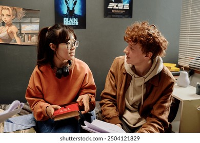 Two students sitting in cozy bedroom setting, discussing study material and holding books. Anime posters on walls and comfortable atmosphere indicating relaxed study environment