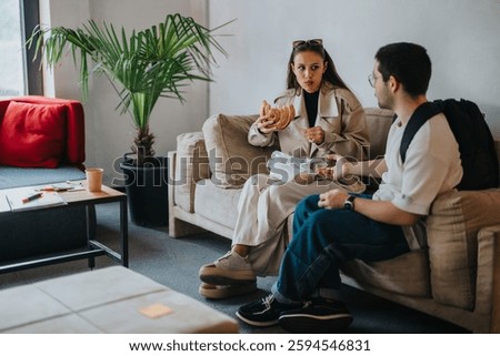 Two students sitting on a sofa, sharing breakfast and conversation during a school break in a cozy lounge area.