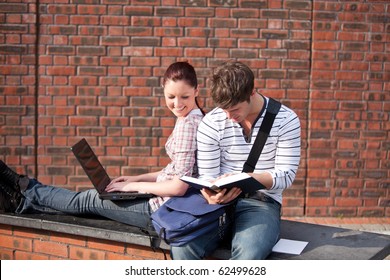 Two Students Working Together With Book And Laptop Outside In The Campus Of Their University