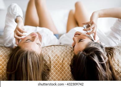 Two Stunning Women Talking On Phones With Their Hair Falling Off The Bed As They Lie Upside Down