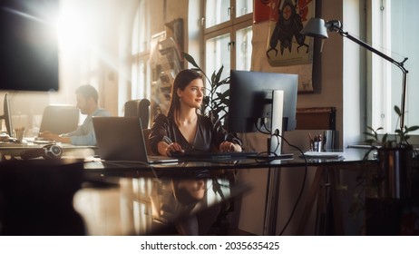 Two Stylish Employees Working on Computers in Creative Agency in Loft Office. Beautiful Manager Typing Correspondence Emails. Sunny Renovated Space with Plants, Artistic Posters and Big Windows. - Shutterstock ID 2035635425
