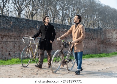 Two stylish men stand with their vintage bicycles, engaging in a lively conversation on a sunlit path with old brick walls and bare trees in the background - Powered by Shutterstock