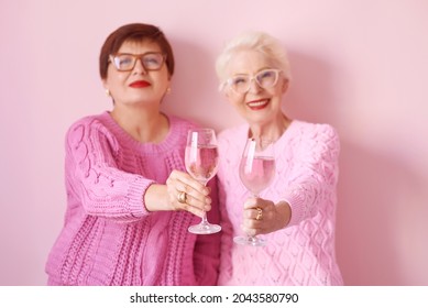 Two Stylish Senior Women In Pink Sweaters With Rose Wine Glasses On Pink Background. Friendship, Talk, Gossip, Relationships, News, Family Concept 