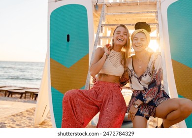 two stylish smiling beautiful women relaxing having fun on beach in summer fashion trend outfit, colorful apparel boho style, having tropical vacation with paddle surf boards - Powered by Shutterstock