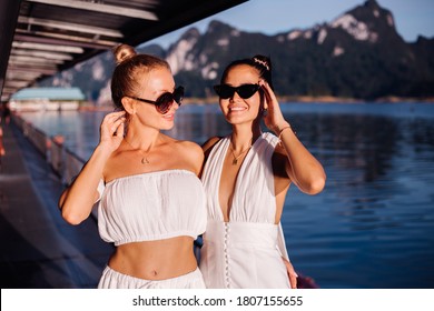 Two Stylish Woman In White Summer Clothes, Dress, Crop Top And High Waist Pants, Wearing Sunglasses. Amazing View On Mountais And Lake On Background, Sunny Day,  Sunset Natural Light.