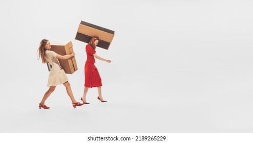 Two Stylish Young Women Carrying Big Cardboard Boxes Isolated Over White Studio Background. Black Friday Shopping, Sales, Moving. Concept Of Retro Fashion, Style, Youth Culture, Emotions, Beauty, Ad