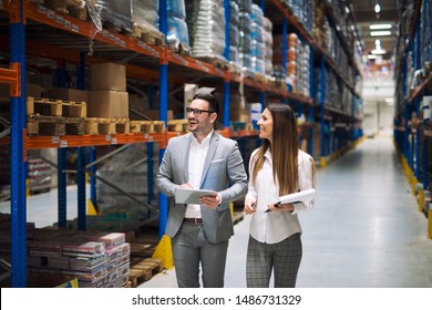 Two successful business people walking through large warehouse center. Manager smiling and looking shelves full with packages and products. Warehouse workers talking about logistics and distribution.