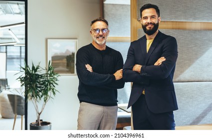 Two successful businessmen smiling at the camera while standing with their arms crossed. Two cheerful businessmen working together in a modern co-working space. - Shutterstock ID 2104396961