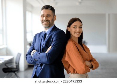 Two successful businesspeople standing back to back with arms crossed and smiling at camera, posing in modern office interior, copy space. Successful businessman and businesswoman