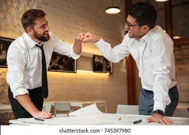 Two successful confident smart businessmen working on a business plan while standing over desk with graph and fist bumping indoors - Powered by Shutterstock