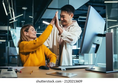 Two successful employees, an Asian man and a woman working in the office at the computer, celebrating victory and success together, clapping their hands - Shutterstock ID 2120213609