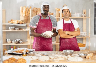 Two successful professional male bakers in maroon aprons posing confidently at floured table in inviting artisan bakery while working with dough - Powered by Shutterstock