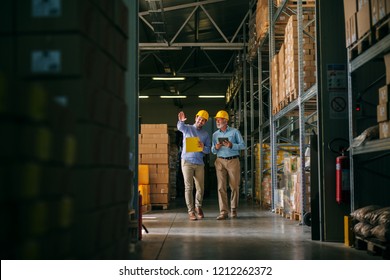 Two Successful Smiling Business Man Walking Through Big Warehouse With Helmets On Their Heads.Younger Man Is Shoving Older One Shelf’s Full Of Products Ready To Be Delivered. Happy Investors.