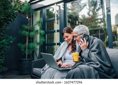 Two successful women of different age sitting in the garden, in blankets, using laptop and talking on smart phone. - Powered by Shutterstock