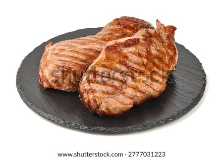 Two succulent, perfectly seared grilled pork chops with rich golden brown crusts and enticing char marks rest on a dark textured slate serving plate, isolated on white background.