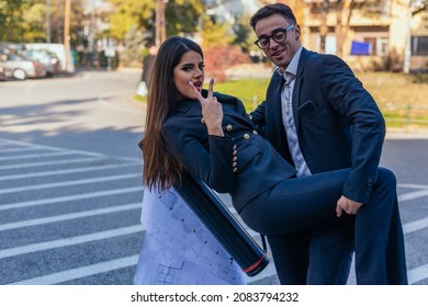 Two suited colleagues posing for the camera. The male colleague is holding the waist and the leg of the female